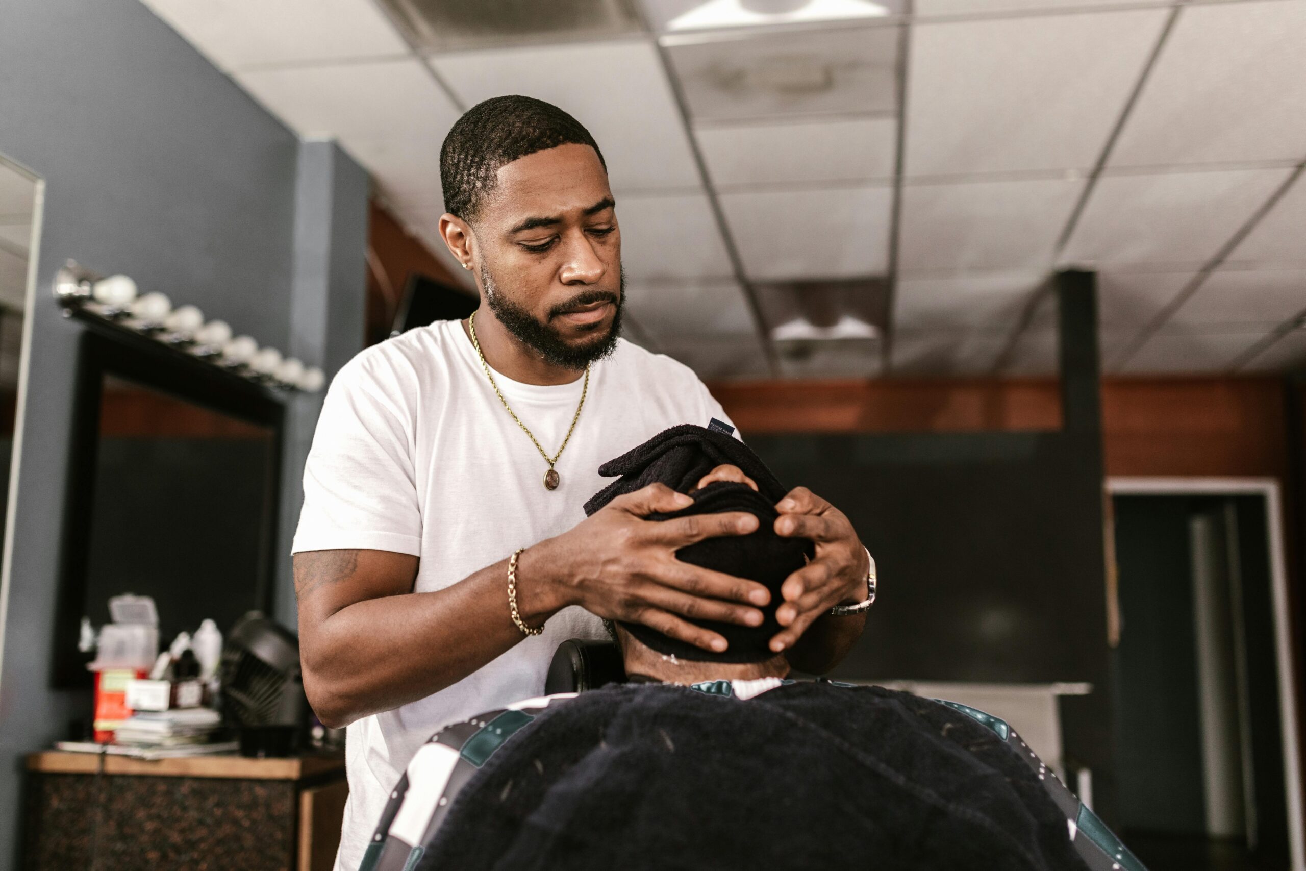 A focused barber provides grooming service to a client with a face towel in a cozy barbershop setting.